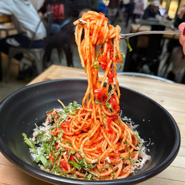 A fork lifts spaghetti with red sauce, basil, and parmesan from a black bowl on a wooden table.