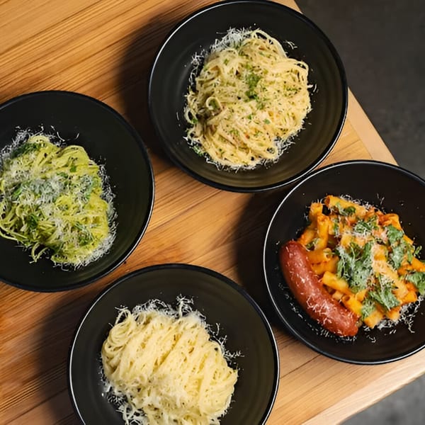 Four black bowls of pasta, including pesto, carbonara, and rigatoni with sausage, on a wooden table.