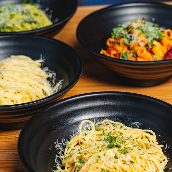 Four black bowls of pasta, including spaghetti with parmesan and parsley, and rigatoni with red sauce.